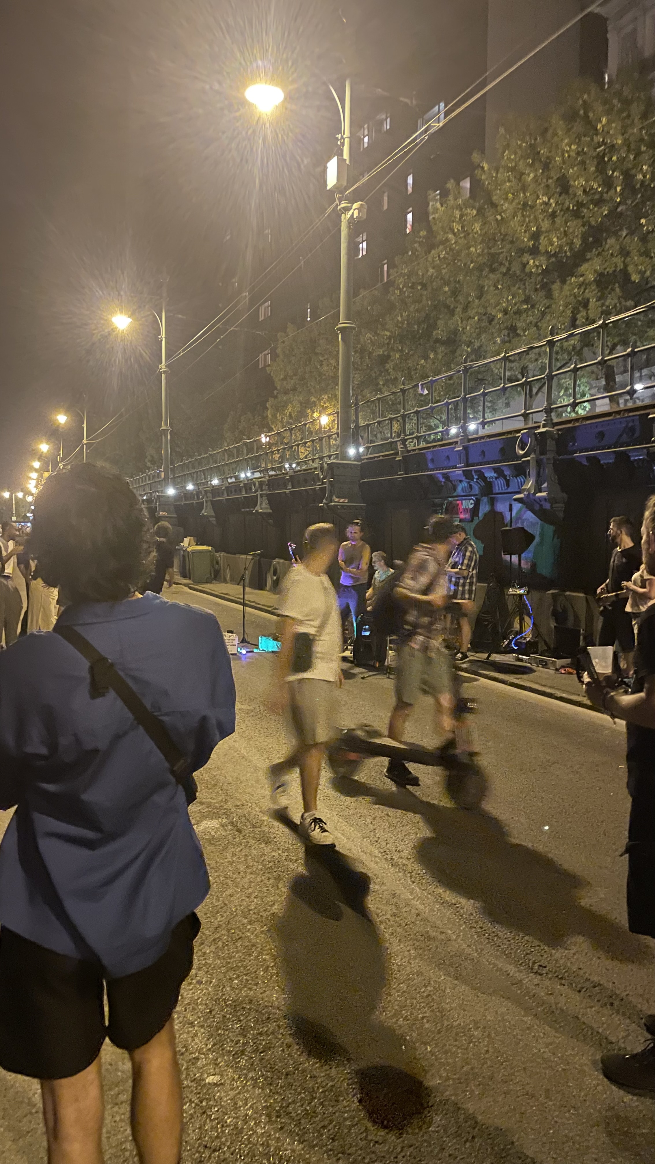 Nighttime street scene by a bridge with streetlights and a crowd gathered around live musicians performing near a railing