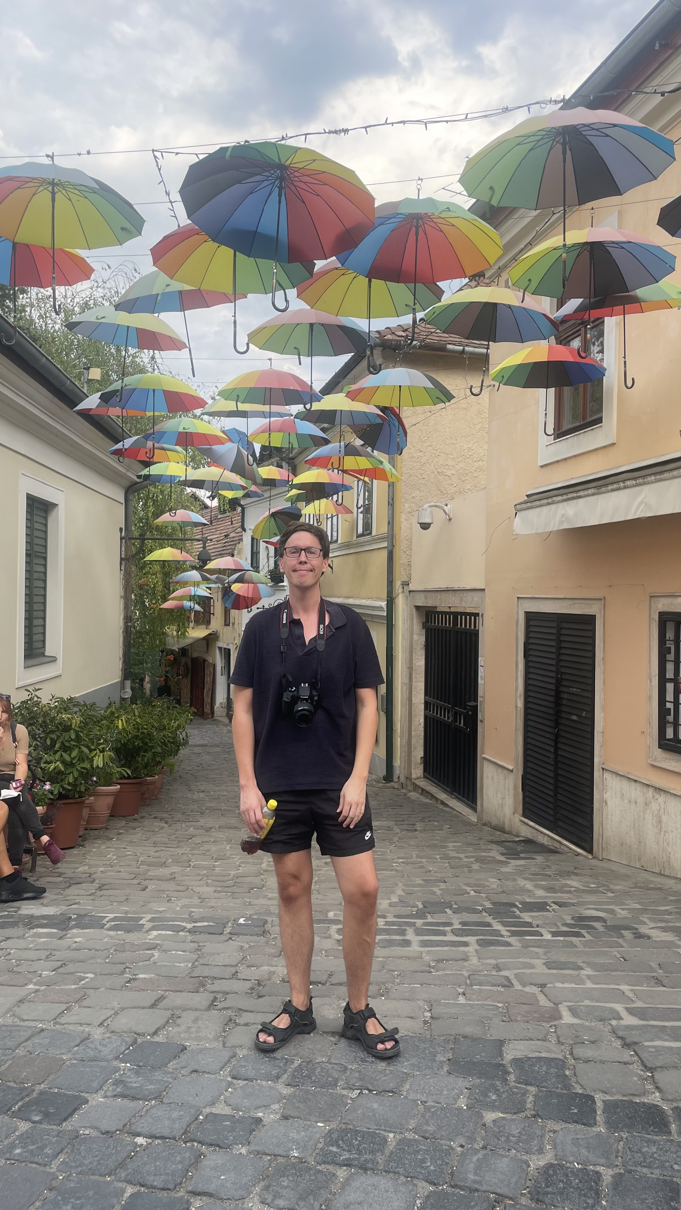 Man standing under a colorful umbrella sky installation on a cobblestone street in Szentendre